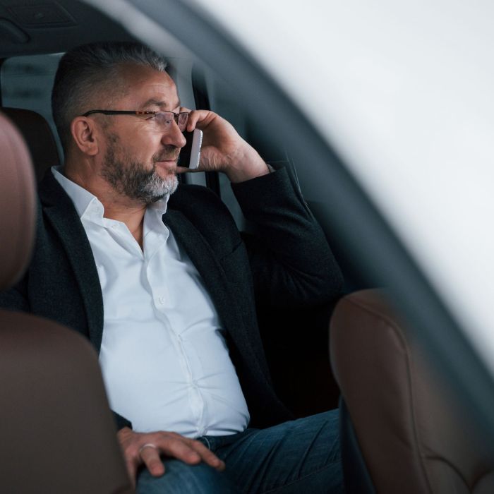 Calm and good mood. Having business call while sitting at the back of modern luxury car. Senior man in glasses and official wear